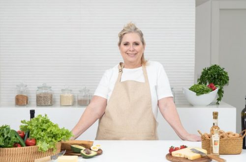 A mature woman in an apron stands at a kitchen counter with fresh produce and cheese, ready for cooking.