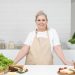A mature woman in an apron stands at a kitchen counter with fresh produce and cheese, ready for cooking.